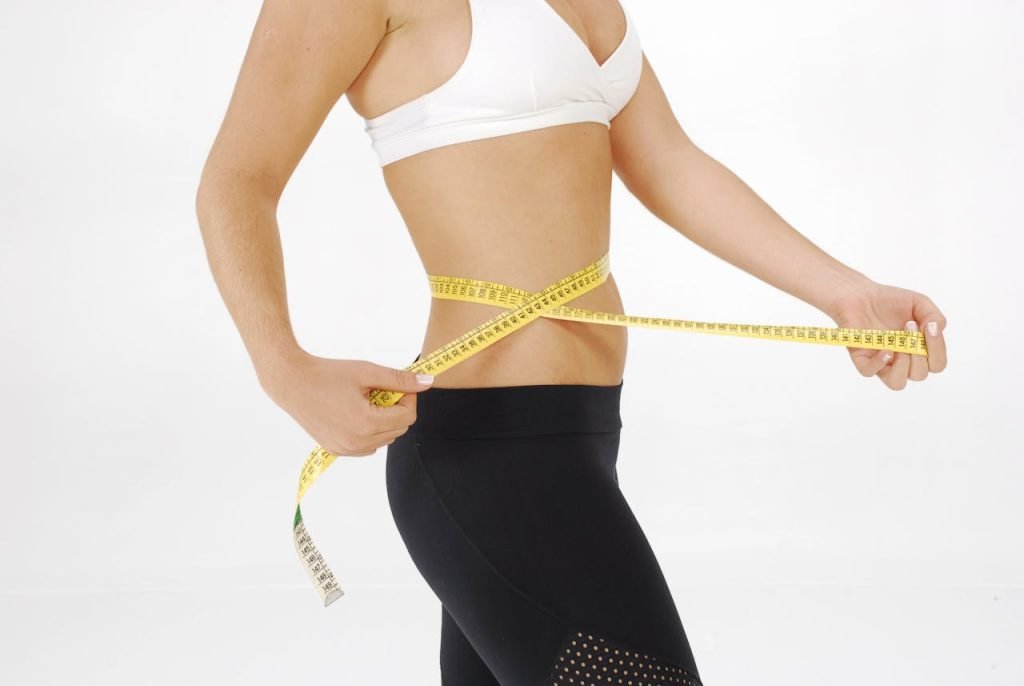 Fit woman in sportswear measuring her waist with a tape measure against a white background.